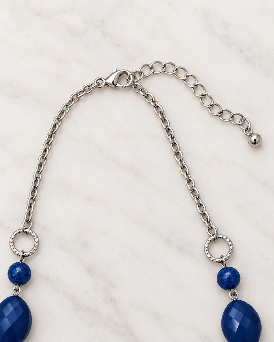 A silver-toned chain necklace with a lobster clasp, adjustable extender chain, textured circular rings, small round beads, and large faceted oval blue gemstone drops, displayed on a white marble surface.