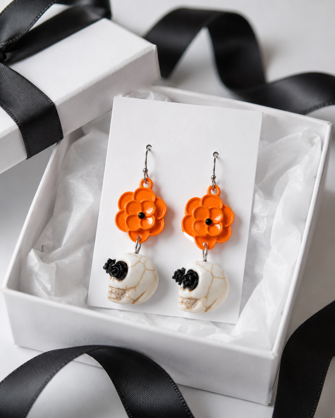 Orange flower and ivory skull earrings displayed on a white jewelry card inside a gift box with black ribbon