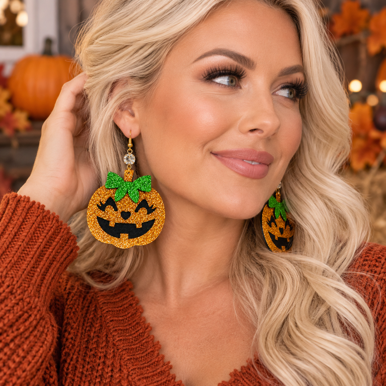 A woman with blonde wavy hair wearing large glittery jack-o'-lantern earrings with green stems, posed in a warm autumn setting with pumpkins in the background.