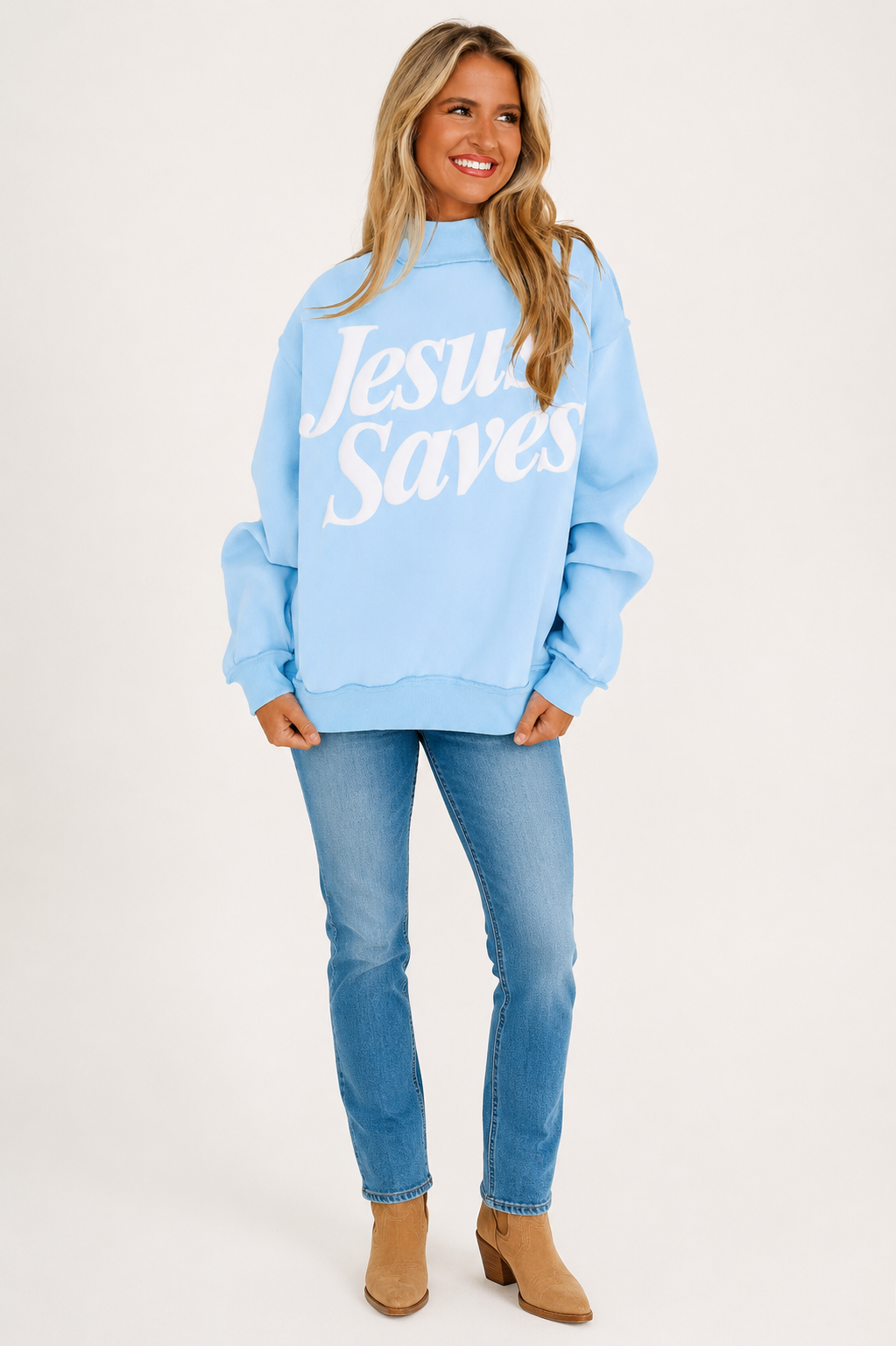 Blonde model wearing the Jesus Saves mist-blue statement sweatshirt with jeans and tan Western boots on a warm white background.