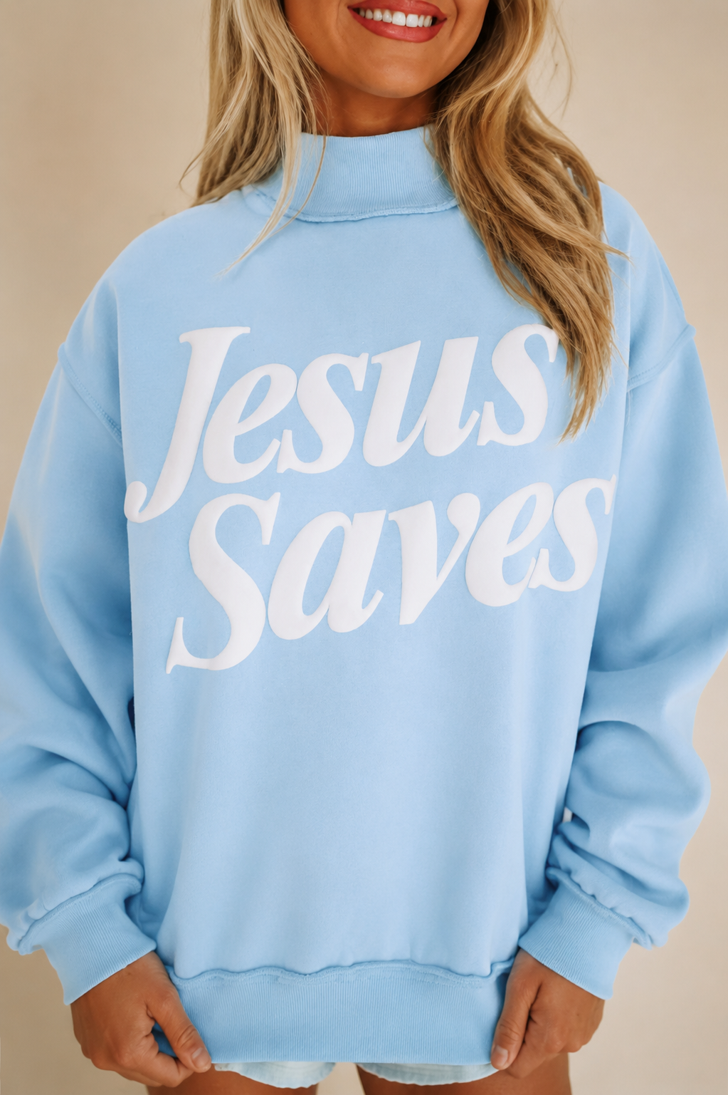 Close view of the mist-blue sweatshirt showing its oversized white Jesus Saves lettering, stand neckline and exposed seam detailing.