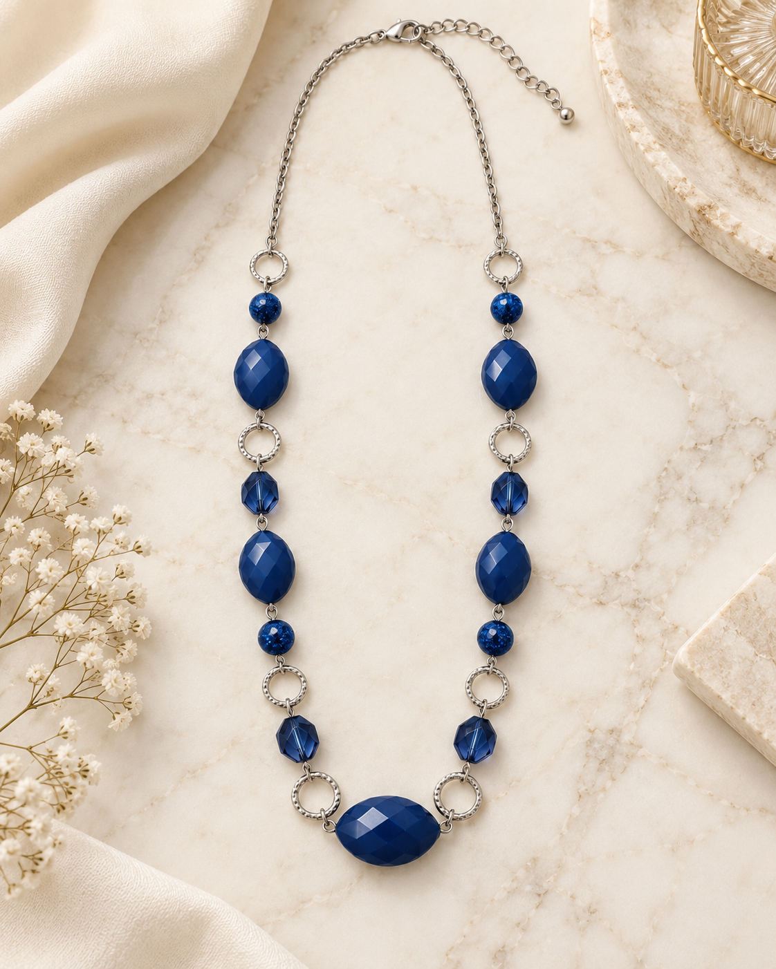 A beaded statement necklace with deep blue faceted beads and silver-toned metal links, displayed on a cream marble surface with neutral fabric and dried flowers.
