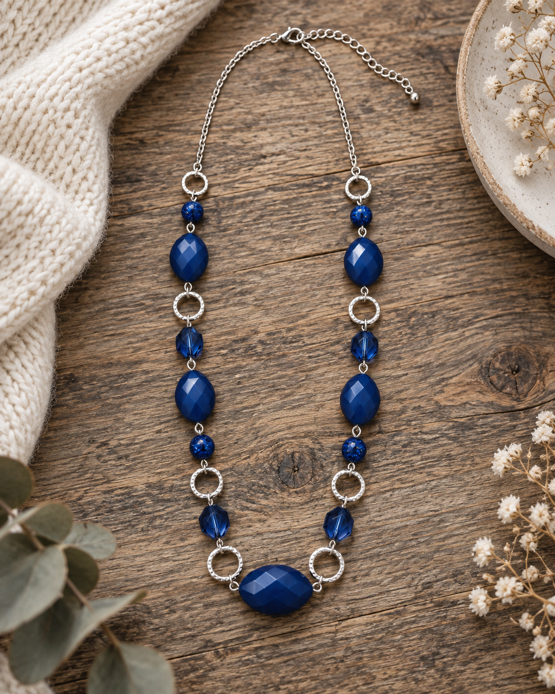 A beaded necklace with deep blue faceted beads and silver-toned metal rings and chain, styled on a rustic wooden surface with a cream knit fabric and dried flowers.