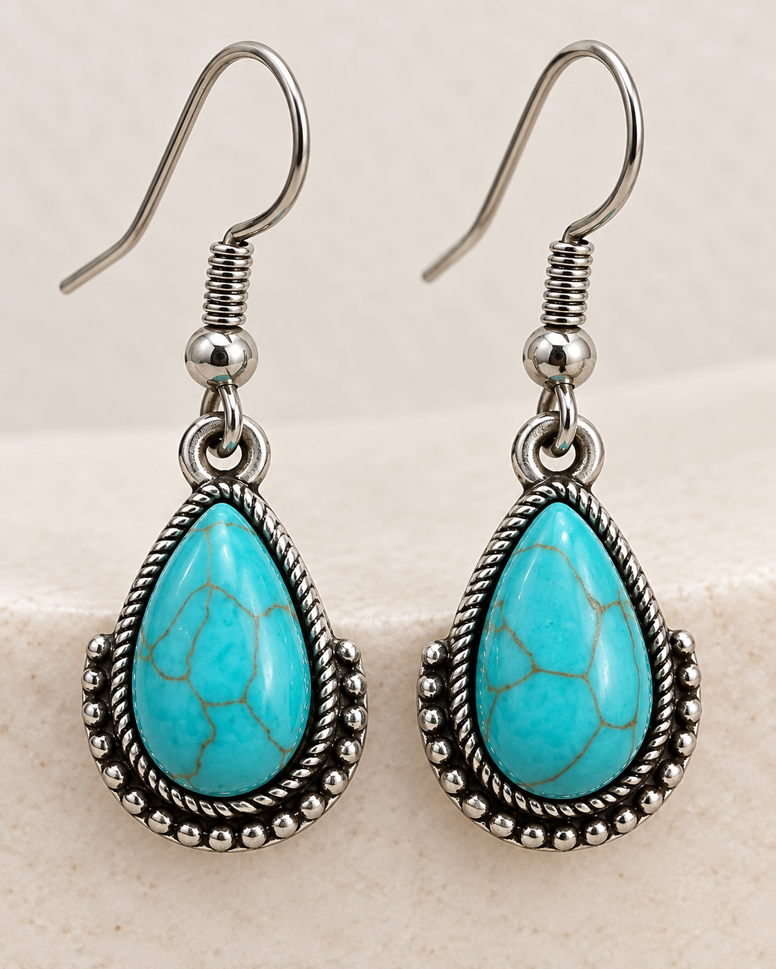 A pair of silver-toned teardrop dangle earrings featuring turquoise-blue marbled stone centers with a rope-textured silver border, displayed on a light beige textured surface.
