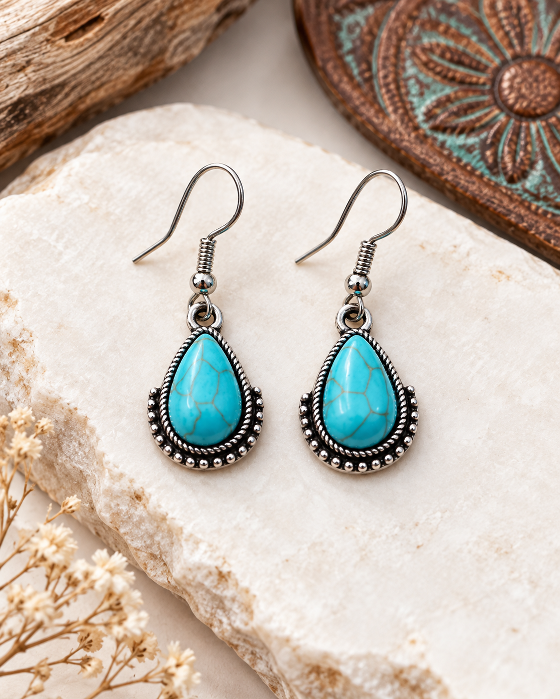 A pair of silver-toned teardrop dangle earrings with vibrant turquoise blue stones, displayed on a textured white stone with rustic dried flowers and wood in the background.