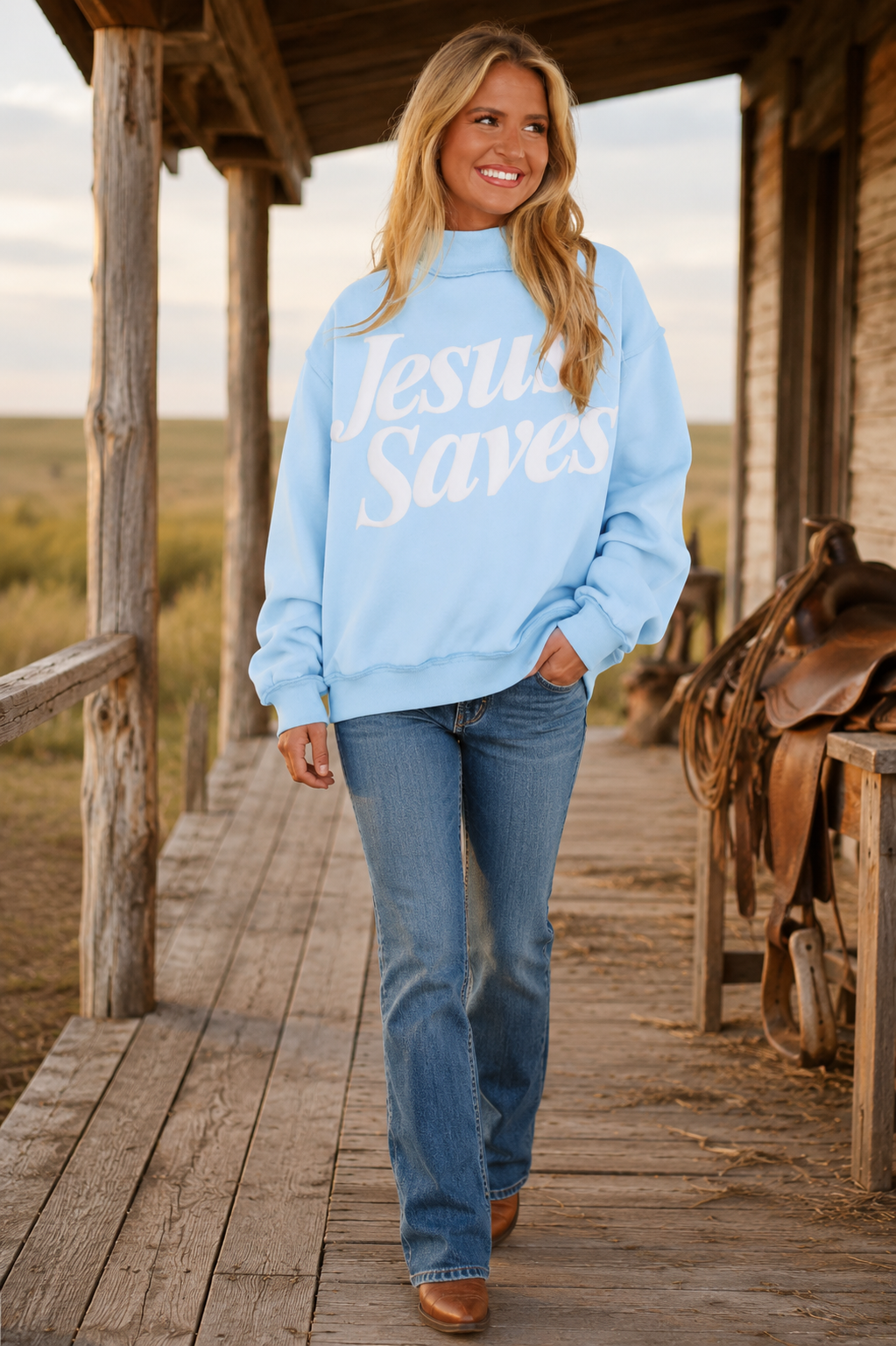 Blonde model wearing the Jesus Saves statement sweatshirt with blue jeans and Western boots on a rustic ranch porch.