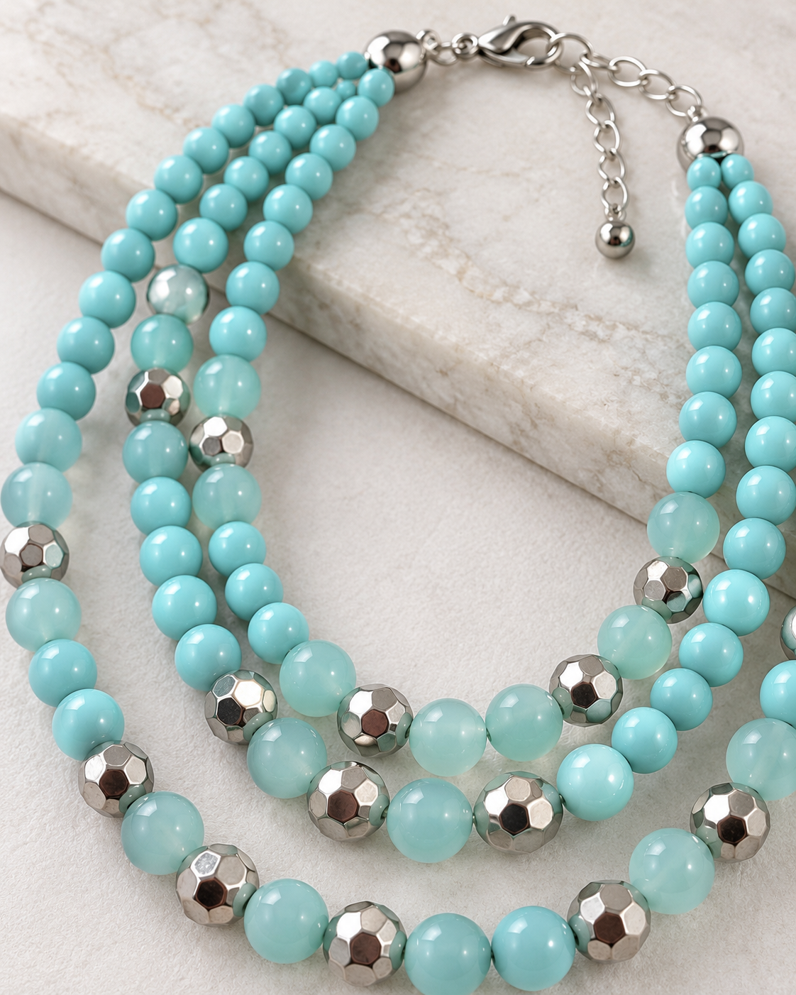 A multi-strand beaded necklace with light aqua blue beads and silver faceted metal accents, displayed on a light marble surface.