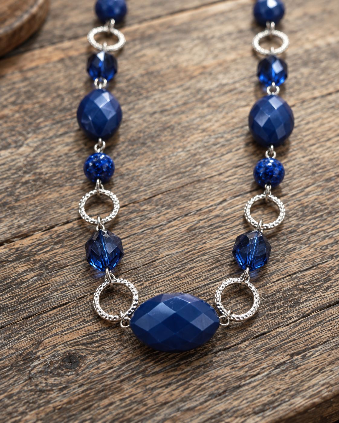 A beaded necklace featuring deep blue faceted beads, textured silver rings, and smaller sparkly blue beads, displayed on a rustic wooden surface.