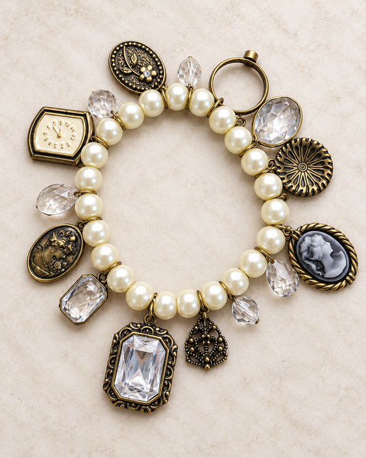 An antique-style charm bracelet with cream pearl beads, bronze-toned metal charms, clear faceted rhinestones, and carved cameo-style medallions, displayed on a light beige textured background.