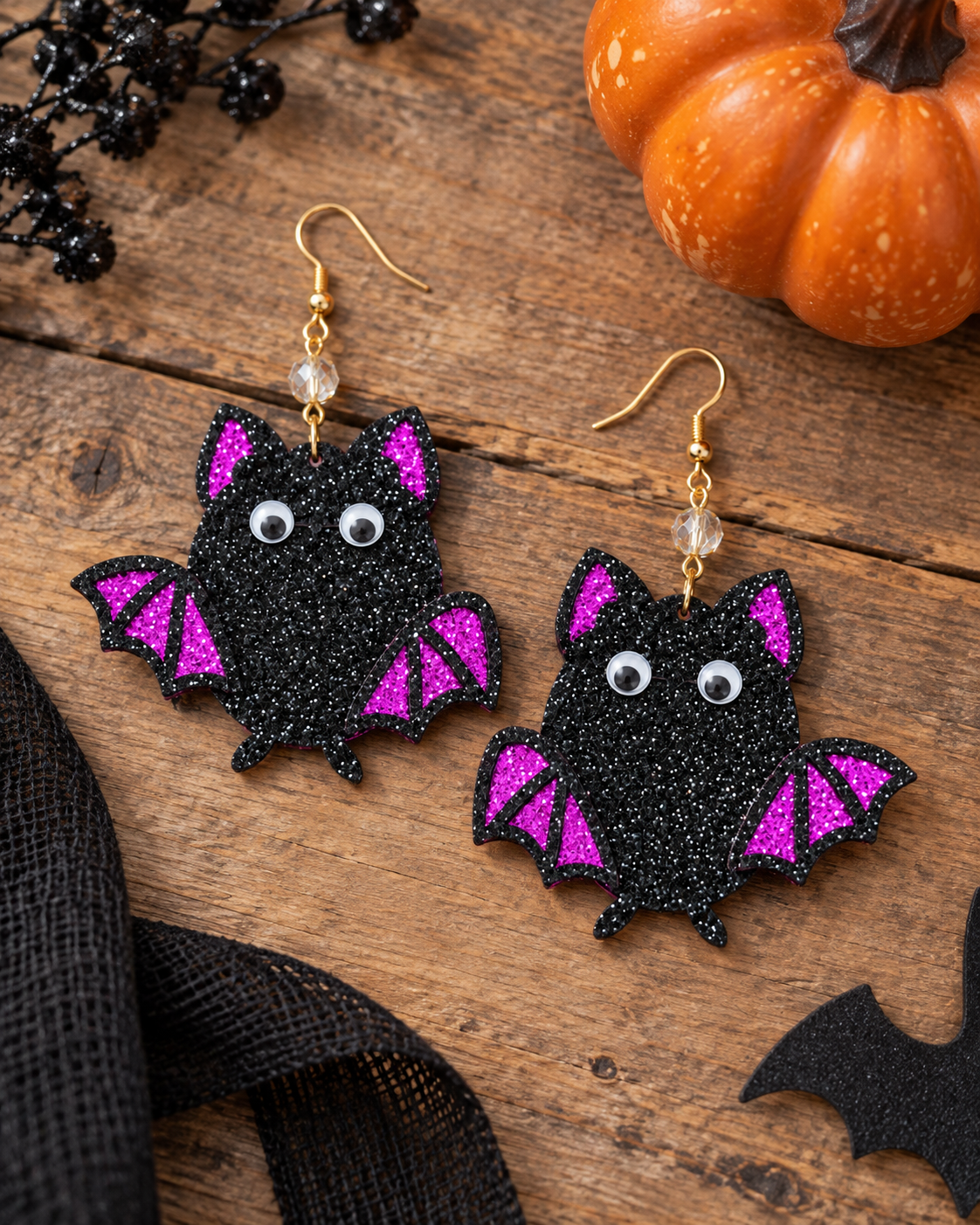 A pair of black glitter bat earrings with bright pink glitter wings and white googly eyes, displayed on a rustic wooden surface with Halloween-themed props including a small pumpkin, black mesh fabric, and black tinsel.