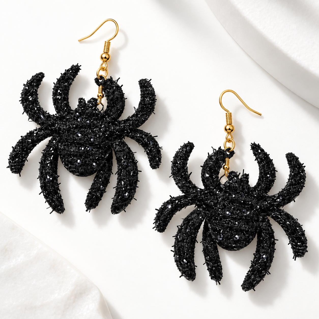 A pair of black spider-shaped dangling earrings with gold fishhook ear wires, photographed on a clean white surface with soft shadows.