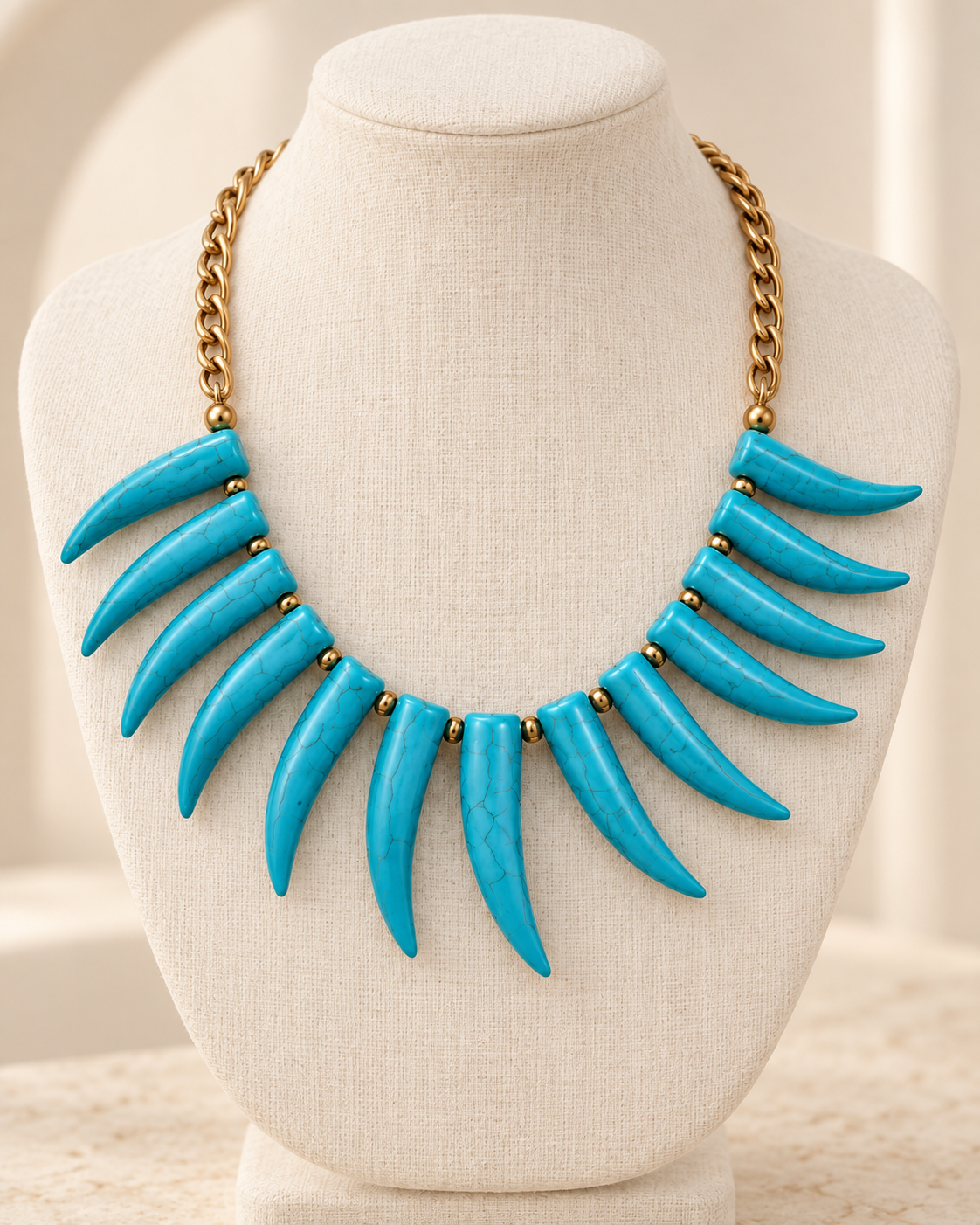 A statement bib necklace with bright turquoise blue claw-shaped elements and a gold-tone chain, displayed on a cream fabric jewelry bust.