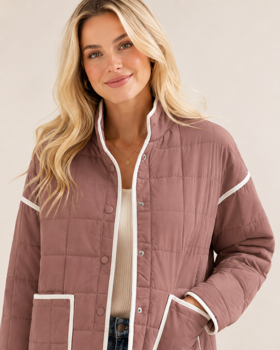 A woman wearing a dusty rose quilted jacket with white contrast trim, layered over a cream ribbed top and blue jeans, against a light beige studio background.