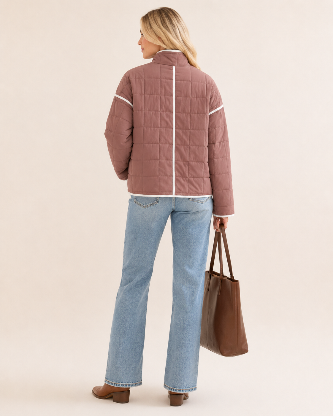 A woman is shown from the back wearing a dusty rose quilted jacket with white piping details, paired with light blue jeans and holding a brown tote bag against a solid beige background.