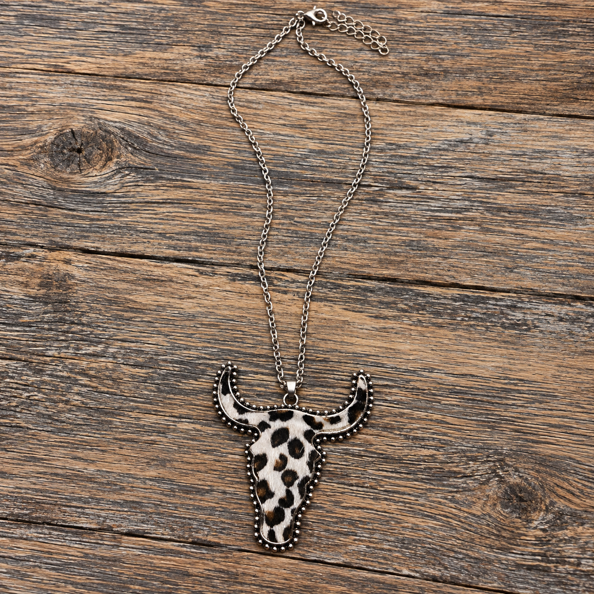 A silver-toned chain necklace with a longhorn skull pendant featuring a black and white spotted pattern, displayed on a rustic wooden surface.