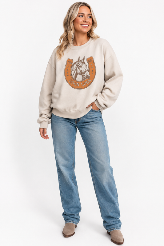 Woman wearing the beige Good Luck horse and horseshoe oversized Western sweatshirt with jeans and boots on a white background.