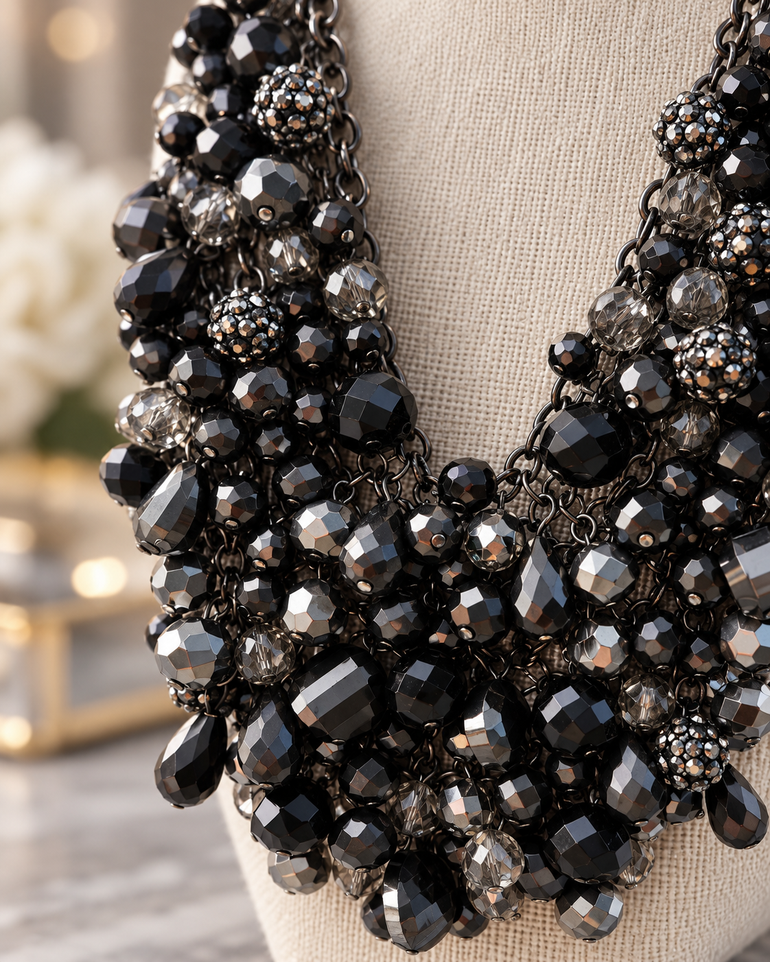 A close-up of a statement bib necklace with layered black faceted beads, teardrop drops, and textured crystal-like accents, displayed on a beige fabric mannequin torso.