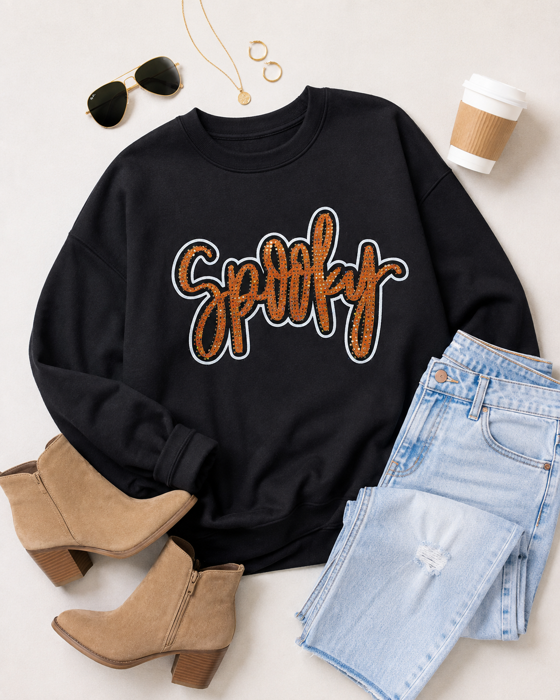 Flat lay of the black Spooky Rhinestone Sweatshirt with light-wash denim, tan ankle boots, gold jewelry, sunglasses, and coffee cup.