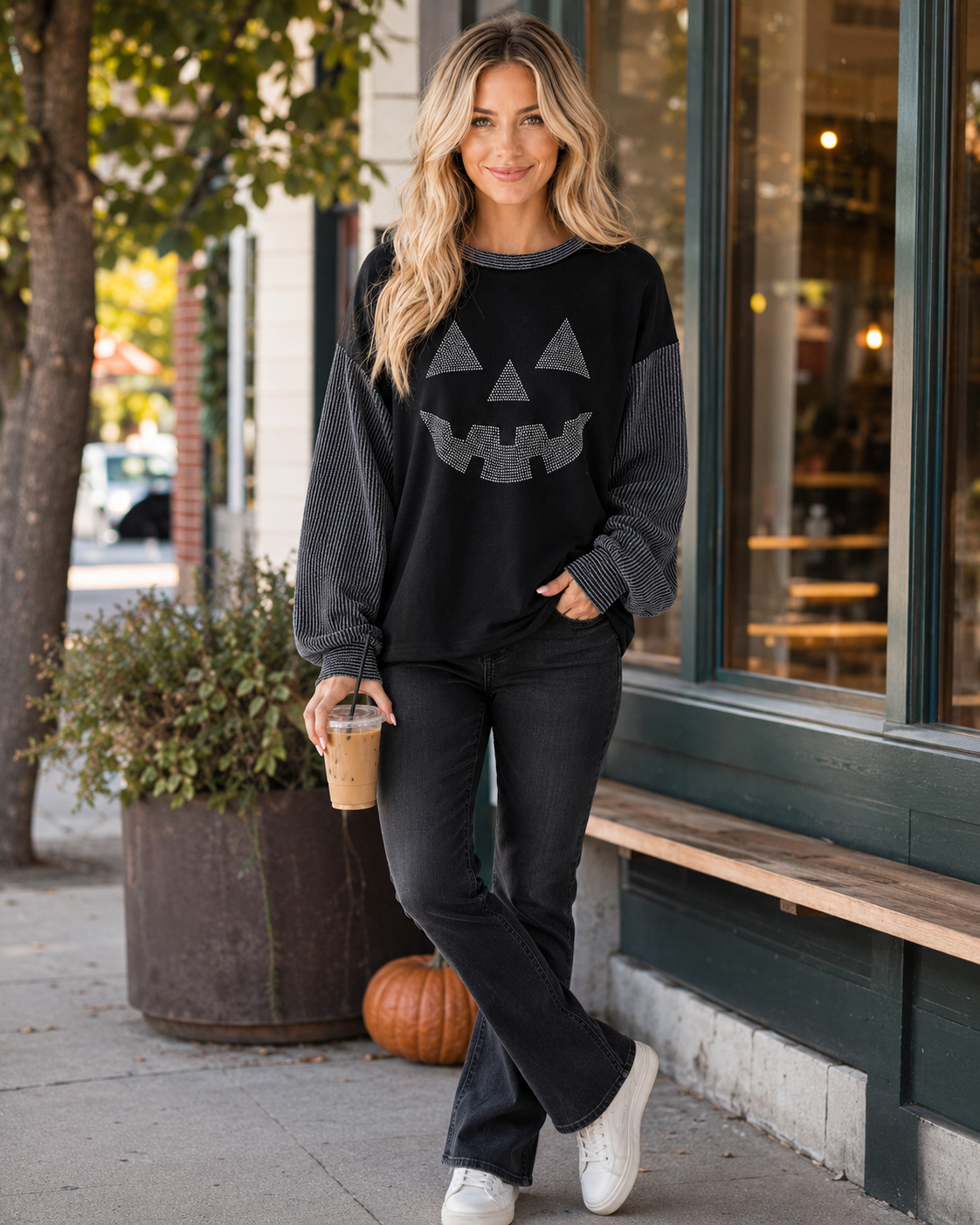 Blonde model styling the black rhinestone pumpkin top with dark denim, white sneakers, and iced coffee outside a café.