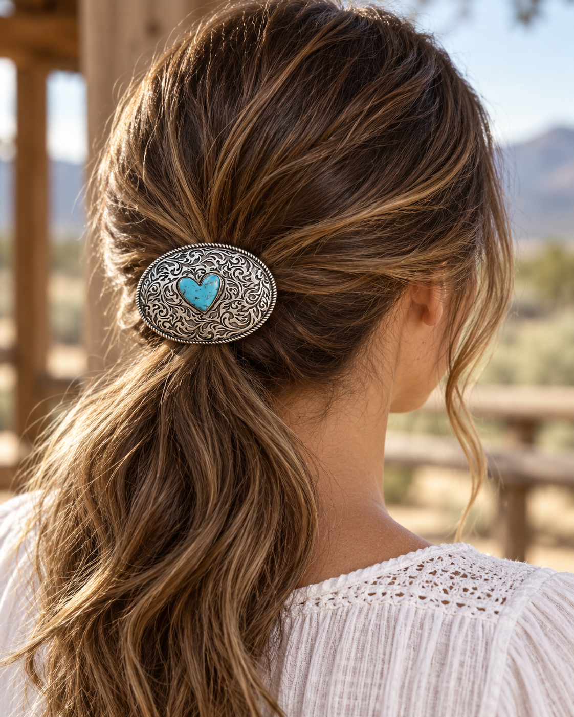 A woman with long brown hair in a ponytail wearing a western-style oval hair clip with an ornate silver filigree border and a turquoise heart in the center, photographed from behind in an outdoor setting.