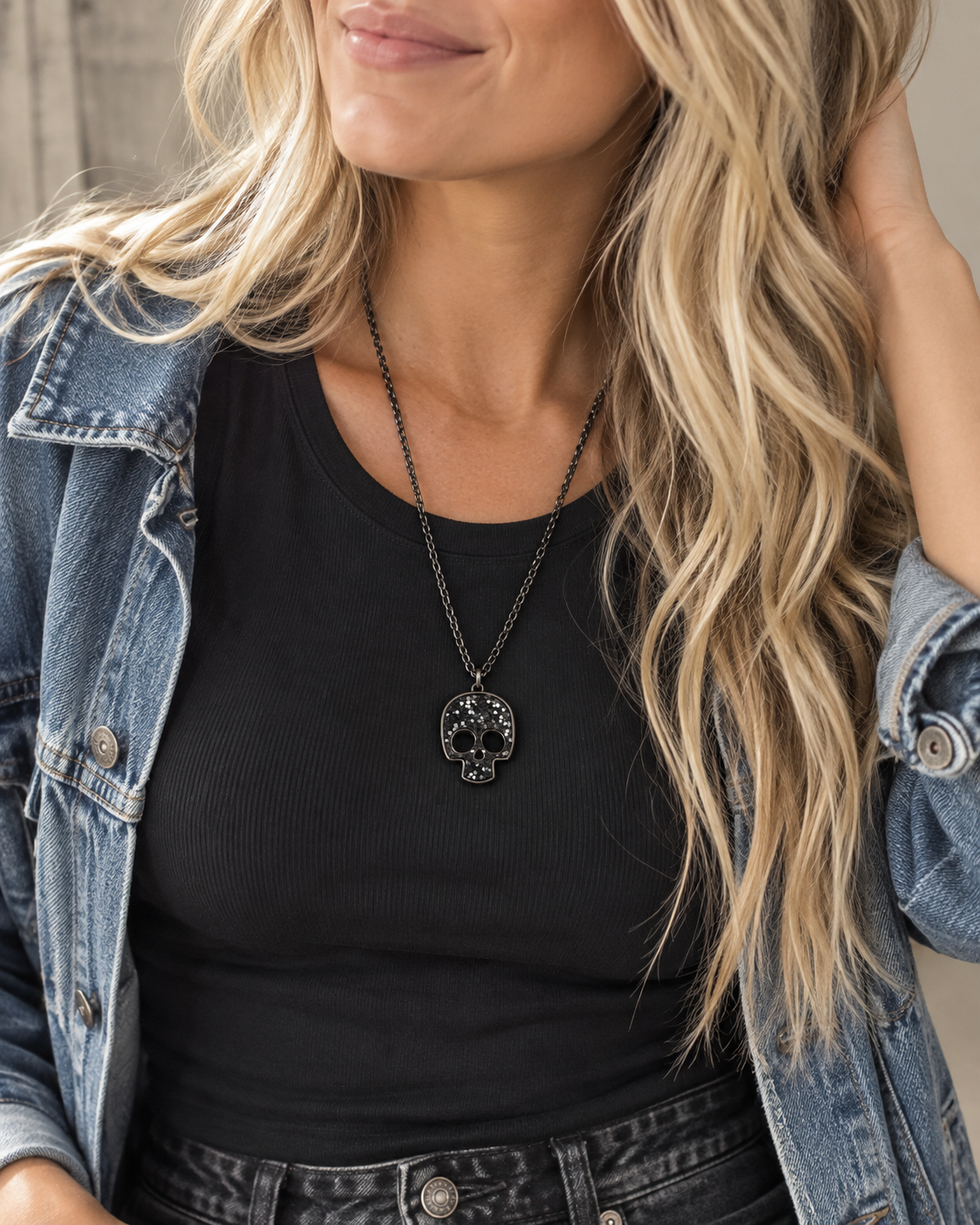 A close-up lifestyle shot of a woman wearing a black pendant necklace with a skull design, styled with a black ribbed tank top and an open blue denim jacket.
