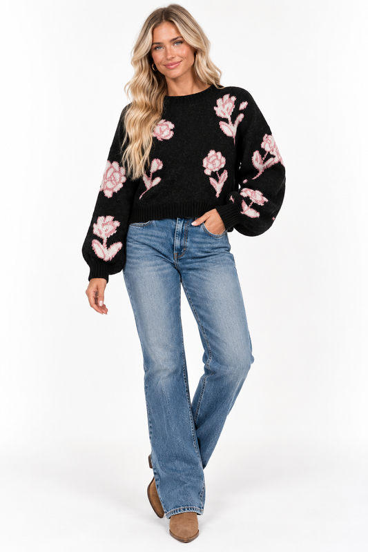 A woman modeling a black knit sweater with pink floral embroidery, paired with blue jeans and brown boots, against a plain white studio background.