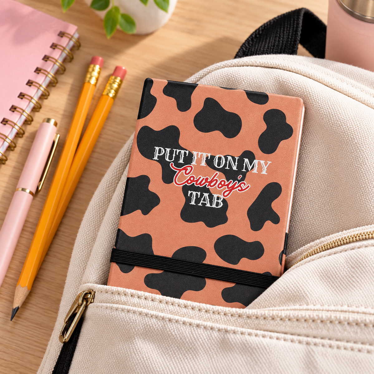 Compact cowprint pocket journal tucked into a cream backpack with pencils and school supplies