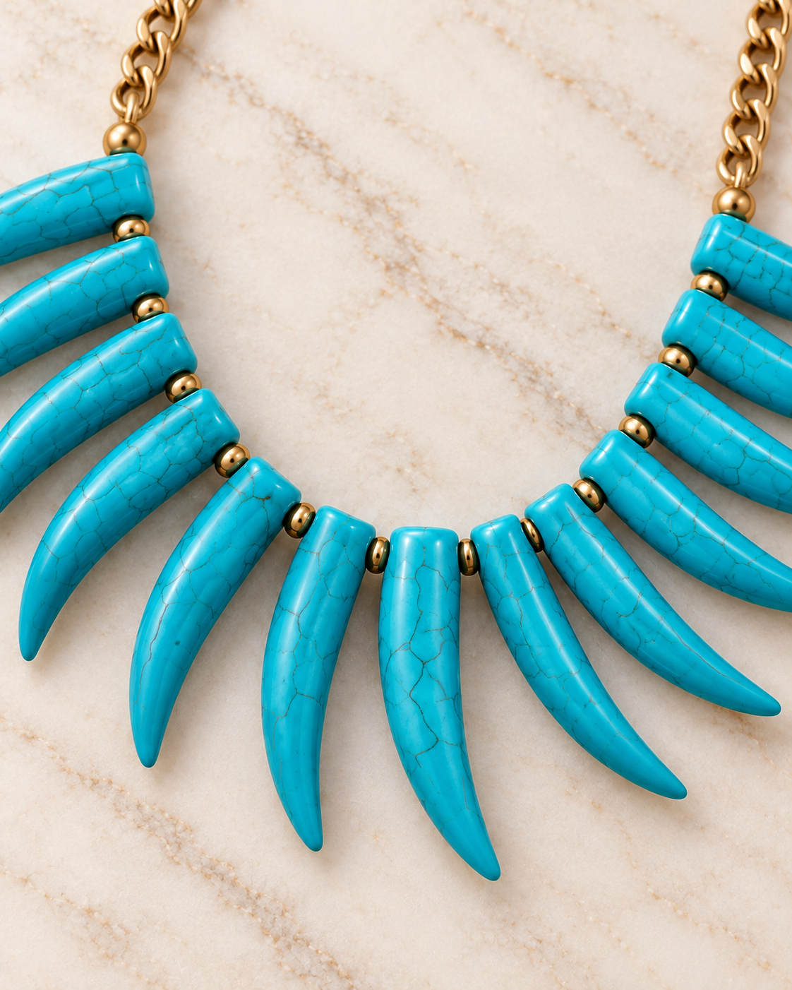 A close-up of a statement bib necklace featuring curved turquoise-colored tooth-like pendants arranged in a fan shape, connected by small gold beads and attached to a gold chain, set against a light beige marble background.