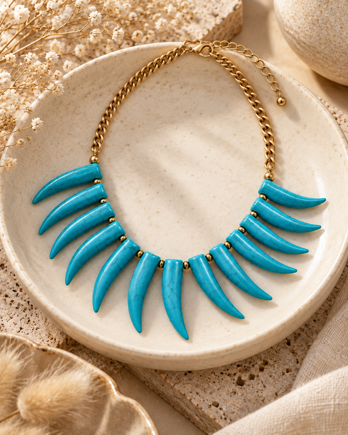 A gold-tone chain necklace with twelve bright turquoise blue claw-shaped teeth, displayed on a round beige ceramic plate with neutral decorative props.