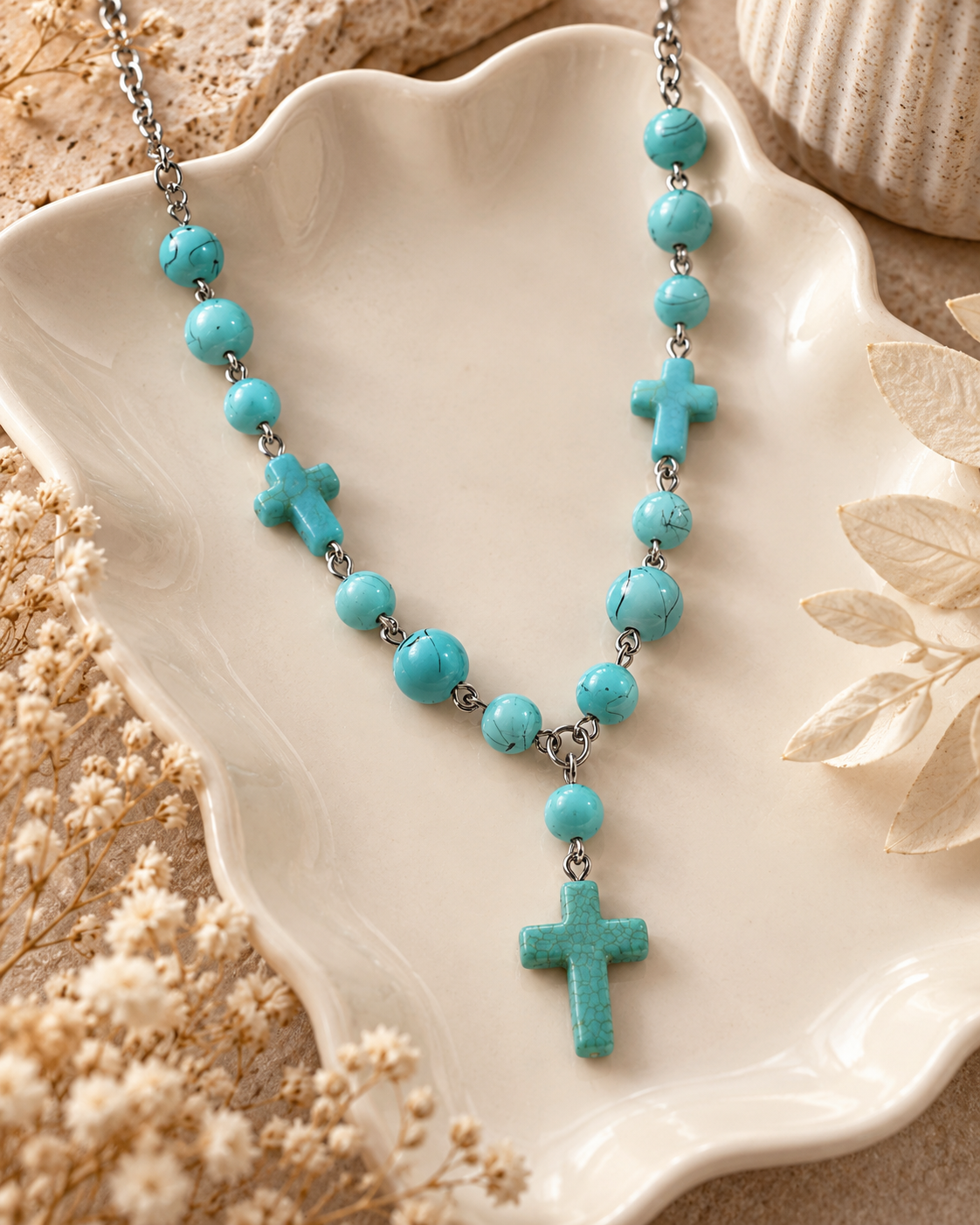 Turquoise-colored cross necklace styled in a cream ceramic dish with soft neutral accessories