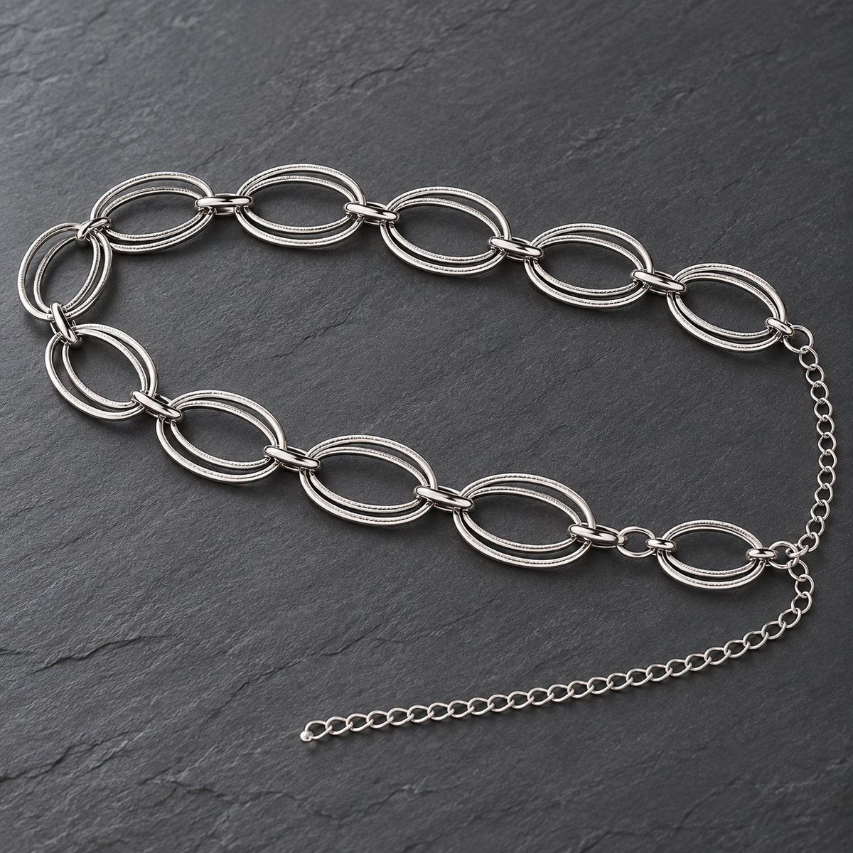 A silver-toned metal chain belt featuring large oval links with a textured finish and an adjustable extension chain, displayed on a dark slate background.