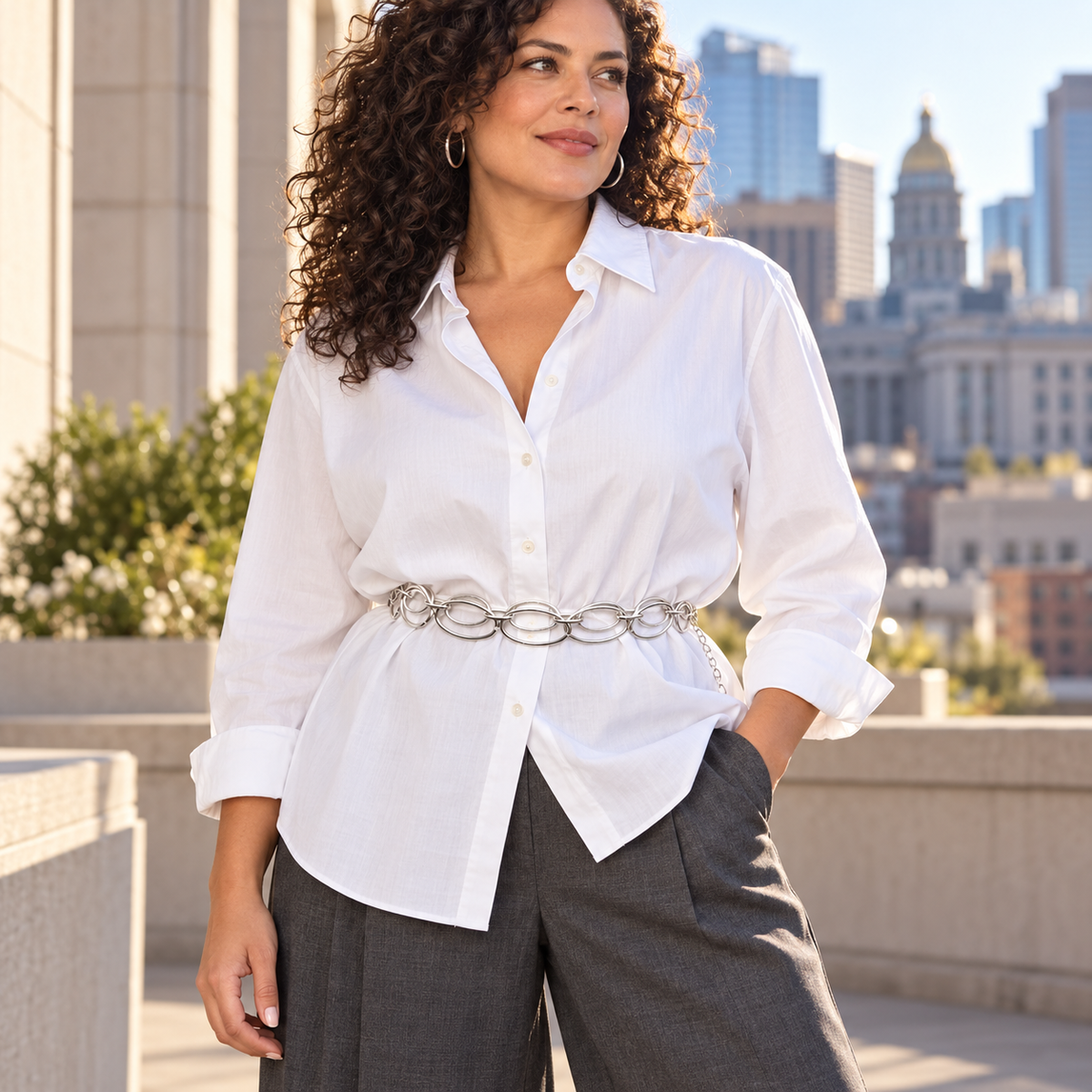 A woman wearing a white button-down shirt tucked into dark gray wide-leg pants, styled with a chunky silver chain belt, standing outdoors on a city balcony.