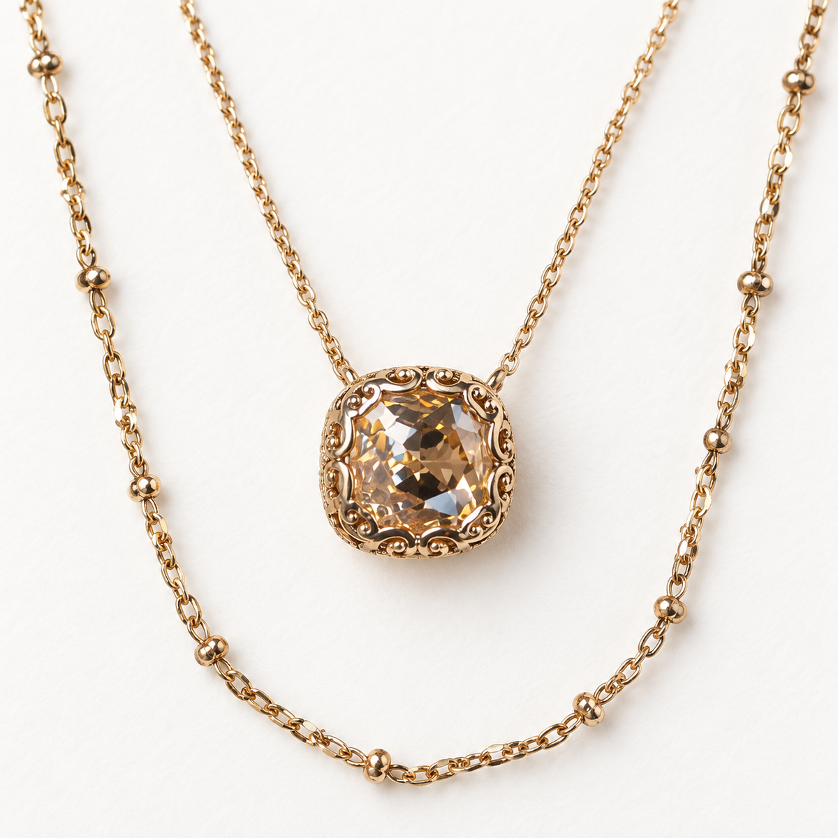 A gold layered necklace featuring a short beaded chain and a longer chain with a square cushion-cut clear gemstone pendant in a detailed gold setting, displayed on a clean white background.