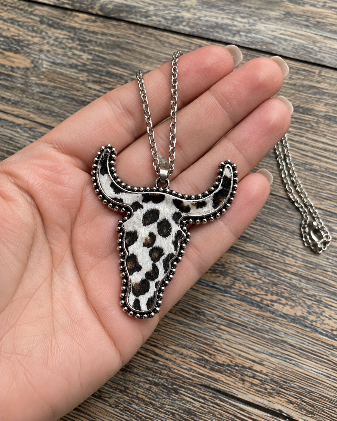 A silver-toned chain necklace with a longhorn-shaped pendant featuring a black, brown, and white spotted animal print, held in a person's hand over a rustic wooden surface.