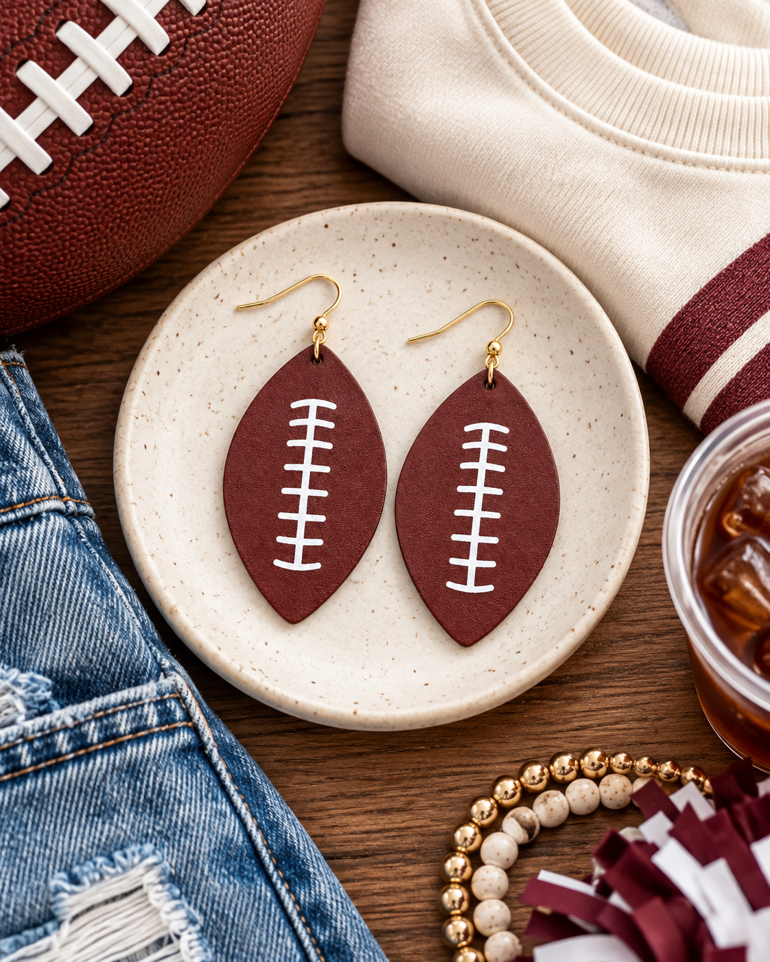 Football-shaped drop earrings styled on rustic wood with gold-tone hooks and white lace details