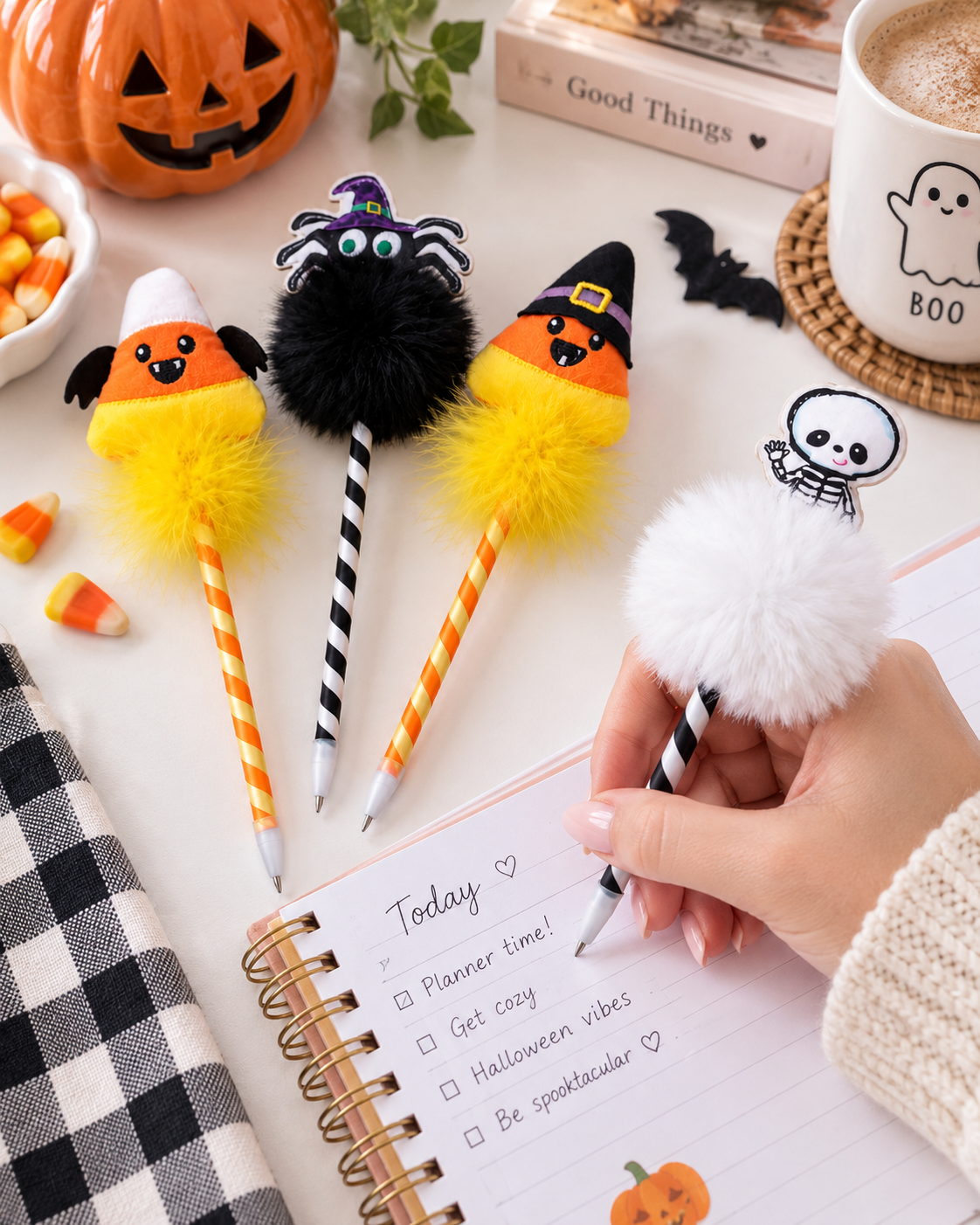Three Halloween-themed decorative pencils with fluffy pom-poms and animal/spooky tops are arranged on a desk beside a hand writing in a notebook. The scene includes candy corn, a jack-o'-lantern, a mug with a ghost and BOO print, and other fall decor, making this a lifestyle product shot.
