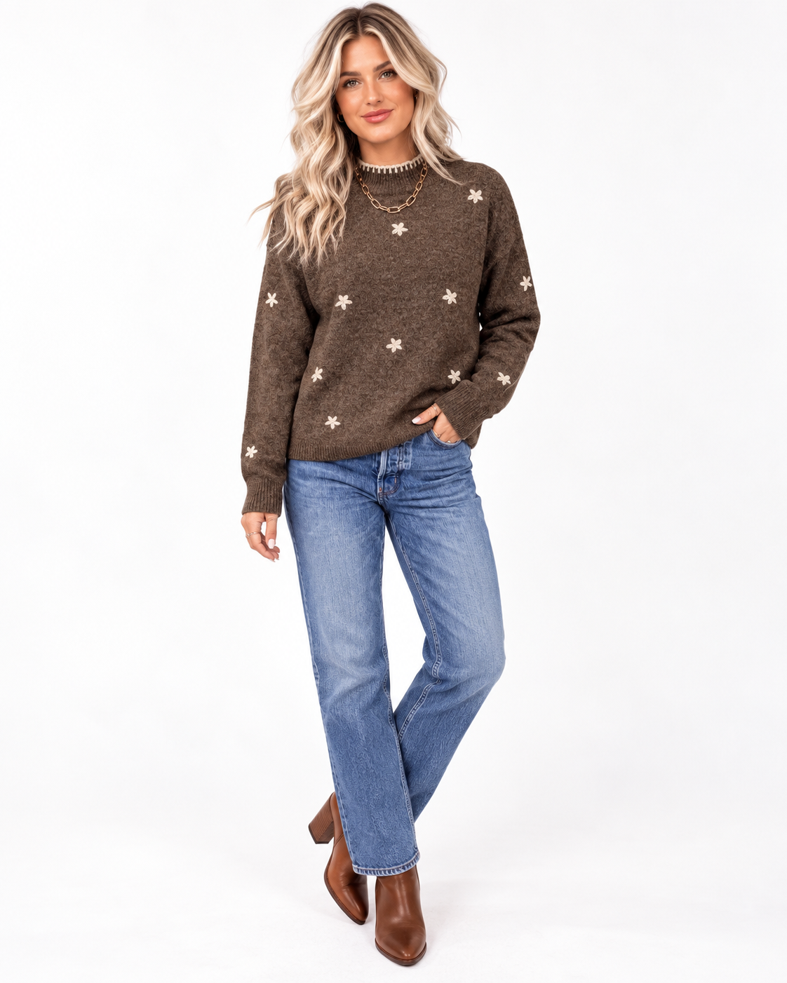 A woman modeling a brown knit sweater with white star patterns, paired with blue jeans and brown ankle boots, against a plain white studio background.