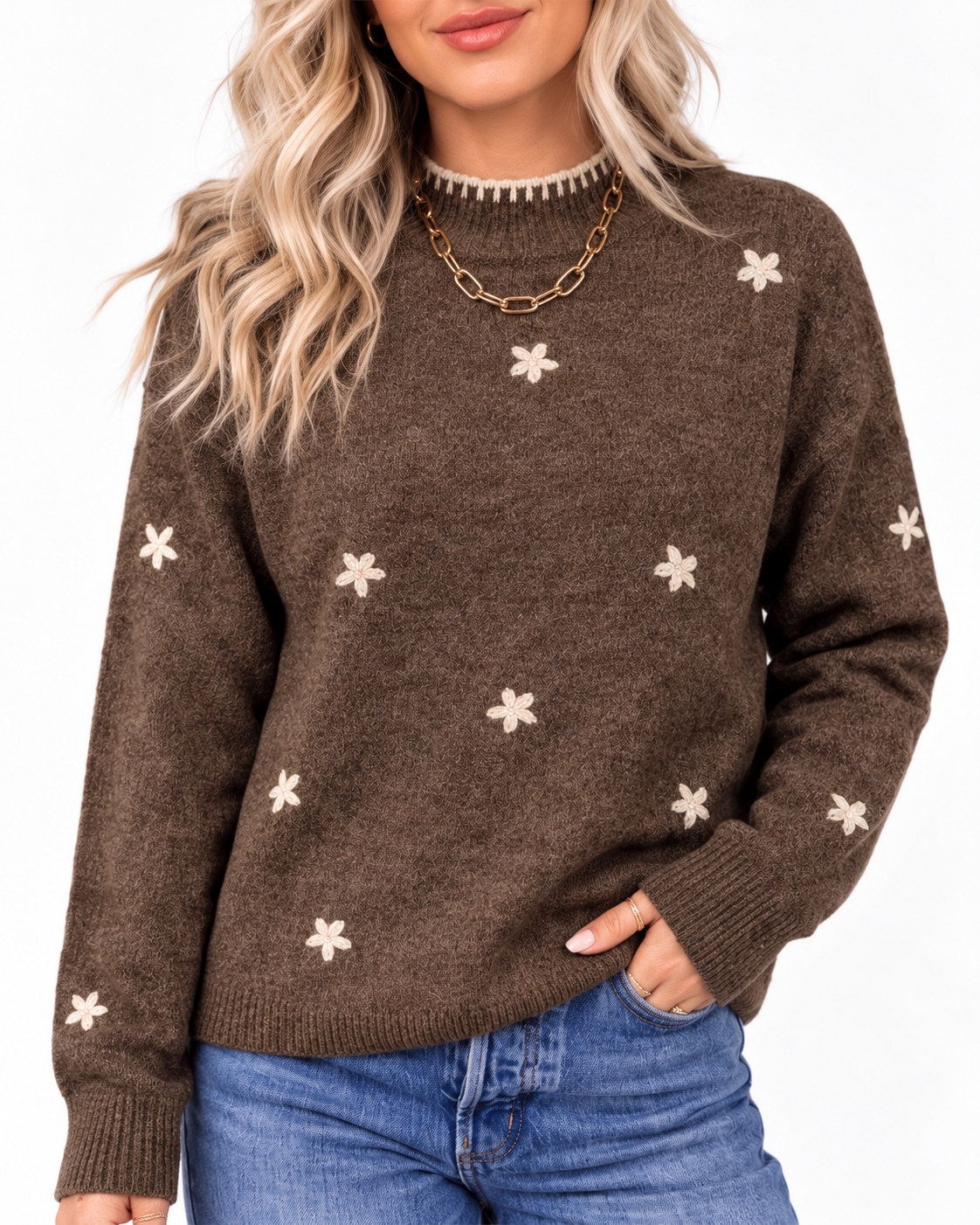 A woman wearing a brown knit sweater with small white floral motifs and a gold chain necklace, paired with blue jeans against a plain white background.