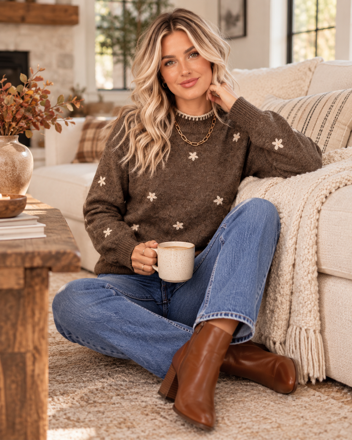 A woman sits cross-legged on a beige rug in a cozy living room, wearing a brown knit sweater with cream star motifs, blue jeans, and brown ankle boots while holding a mug.