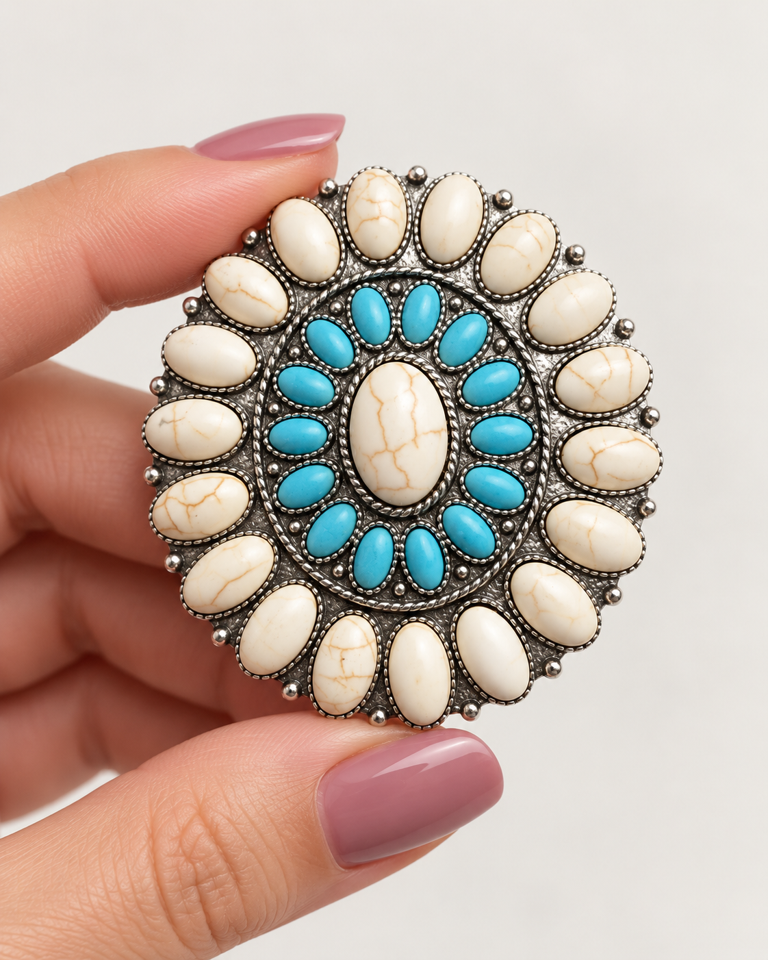 A hand with mauve nail polish holds a round silver-toned brooch featuring a central turquoise and cream stone motif surrounded by a border of cream stones.