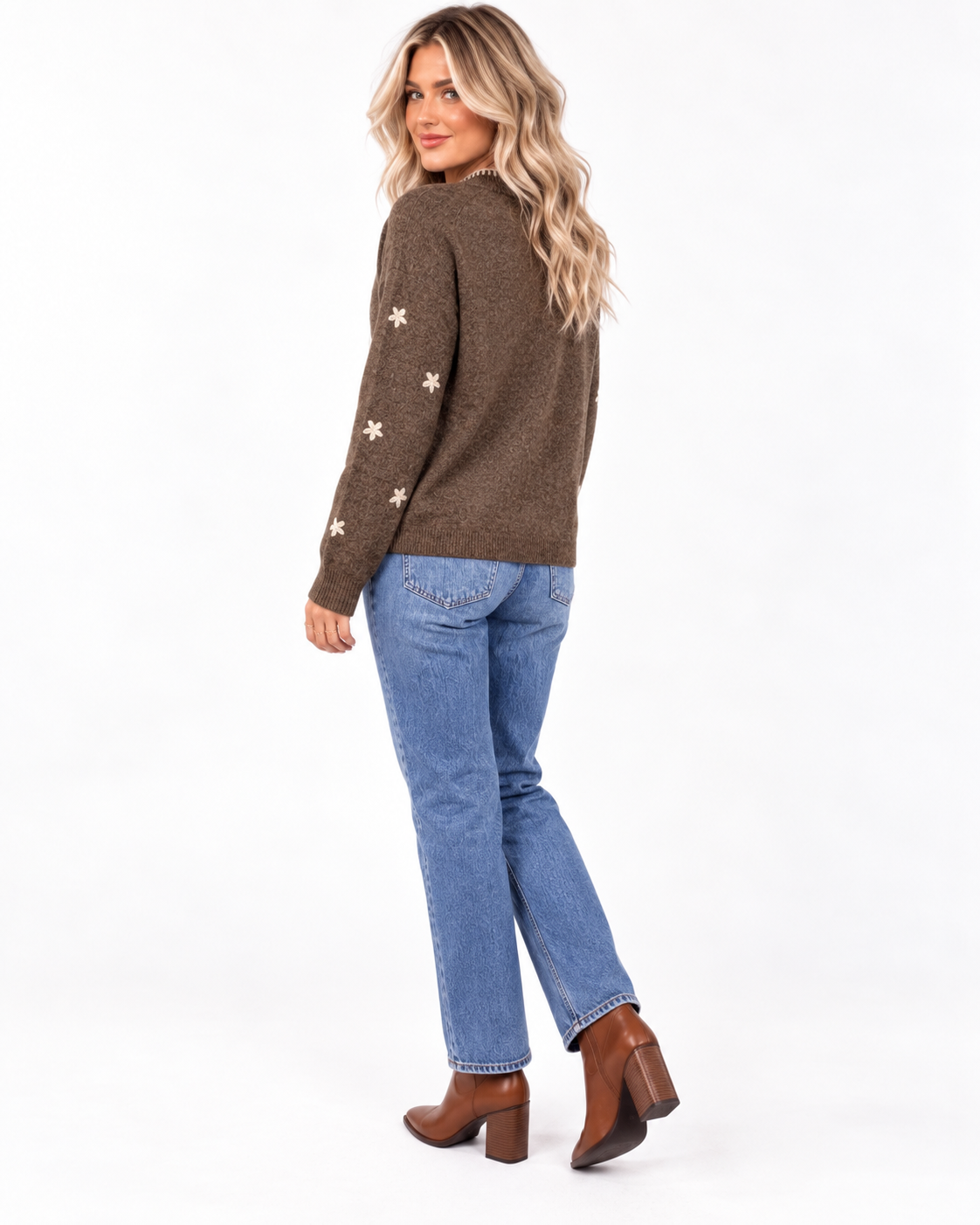 A woman modeling a brown knit sweater with white embroidered star details on the sleeves, paired with blue jeans and brown ankle boots, shown from a three-quarters back view against a white studio background.