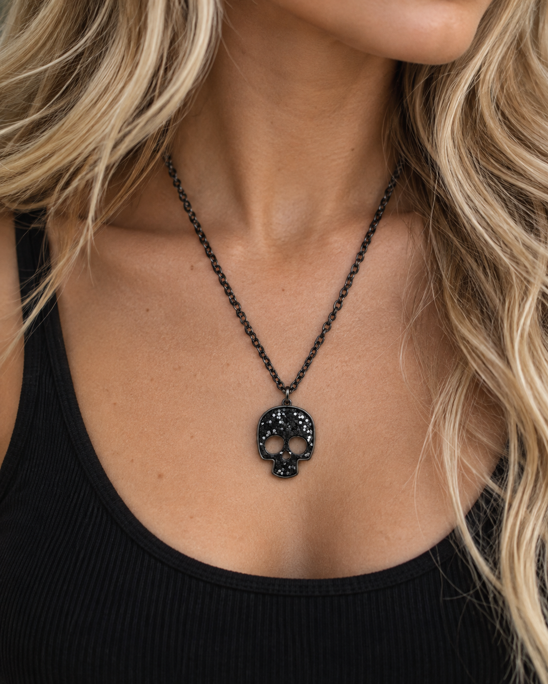 A close-up of a model wearing a black skull pendant necklace on a black ribbed tank top. The necklace features a detailed skull pendant with sparkling clear stones, suspended from a black chain.