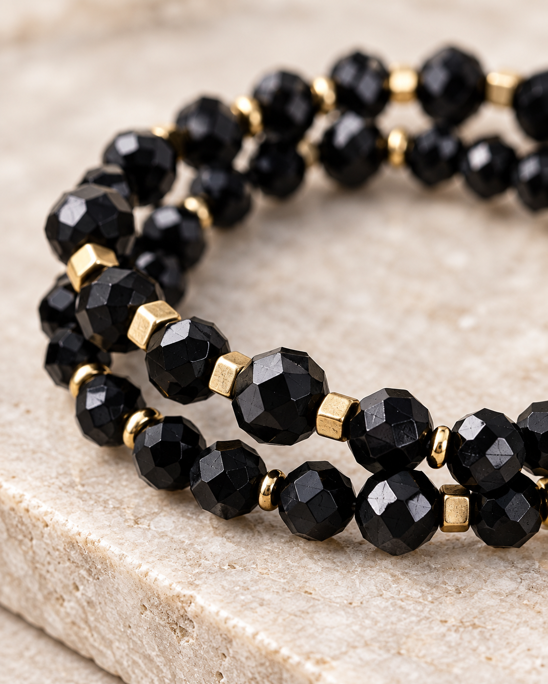 A close-up of a beaded stretch bracelet featuring faceted black beads and small gold spacer cubes, resting on a textured beige stone surface.