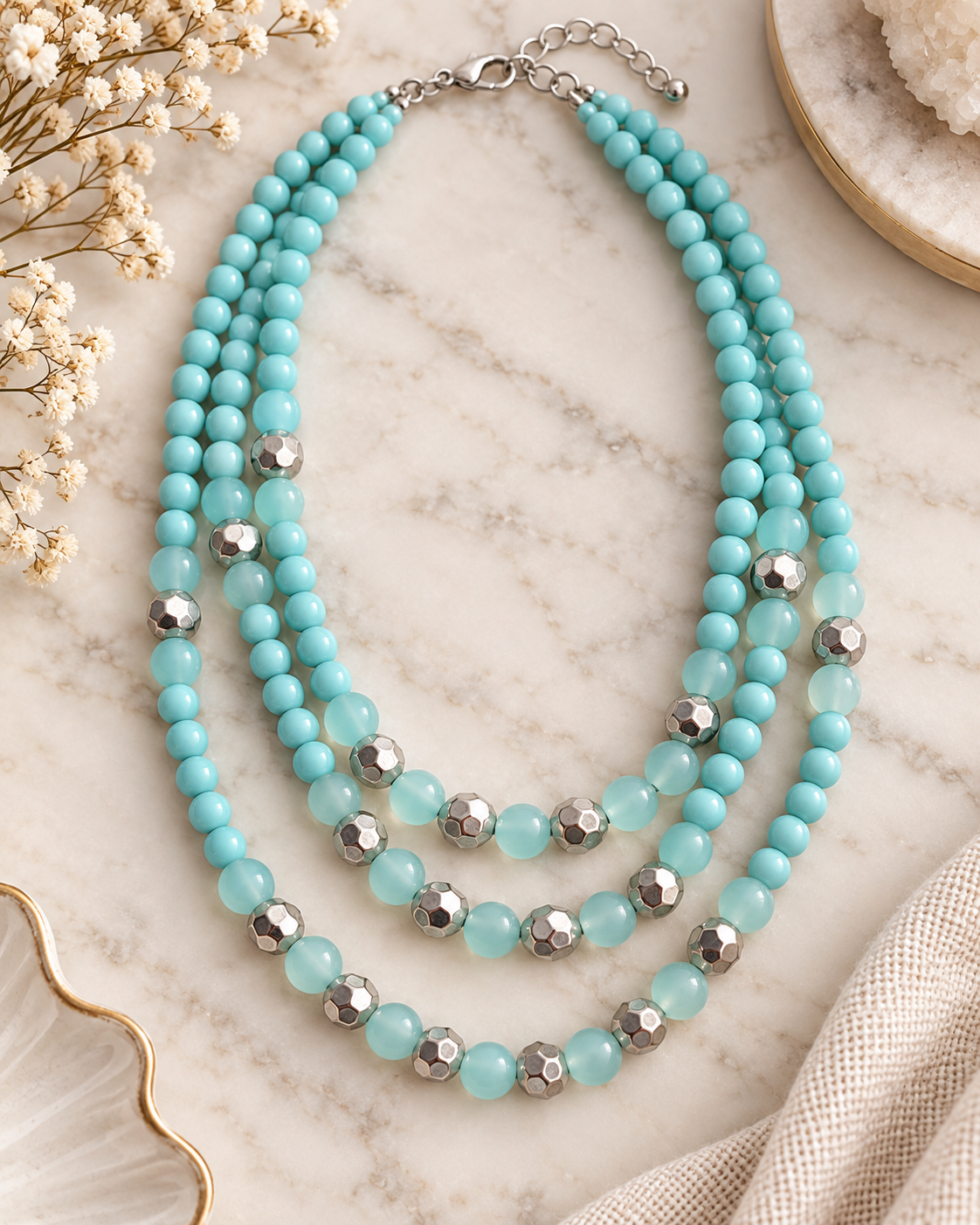 A multi-strand necklace with light turquoise-blue beads and faceted silver metallic accents, styled on a light marble surface with dried flowers and neutral fabric props.