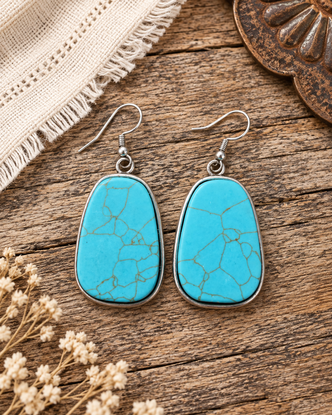 A pair of turquoise stone drop earrings with silver metal borders and fishhook wires, displayed on a rustic wooden surface with dried flowers and a cream woven textile.