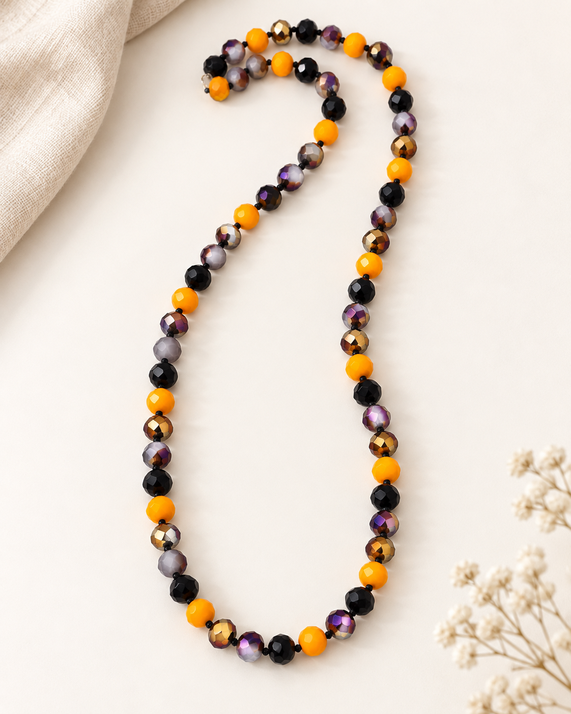 A beaded necklace with alternating bright orange and glossy black faceted beads, laid out on a light cream background with a beige cloth and dried flowers as props.