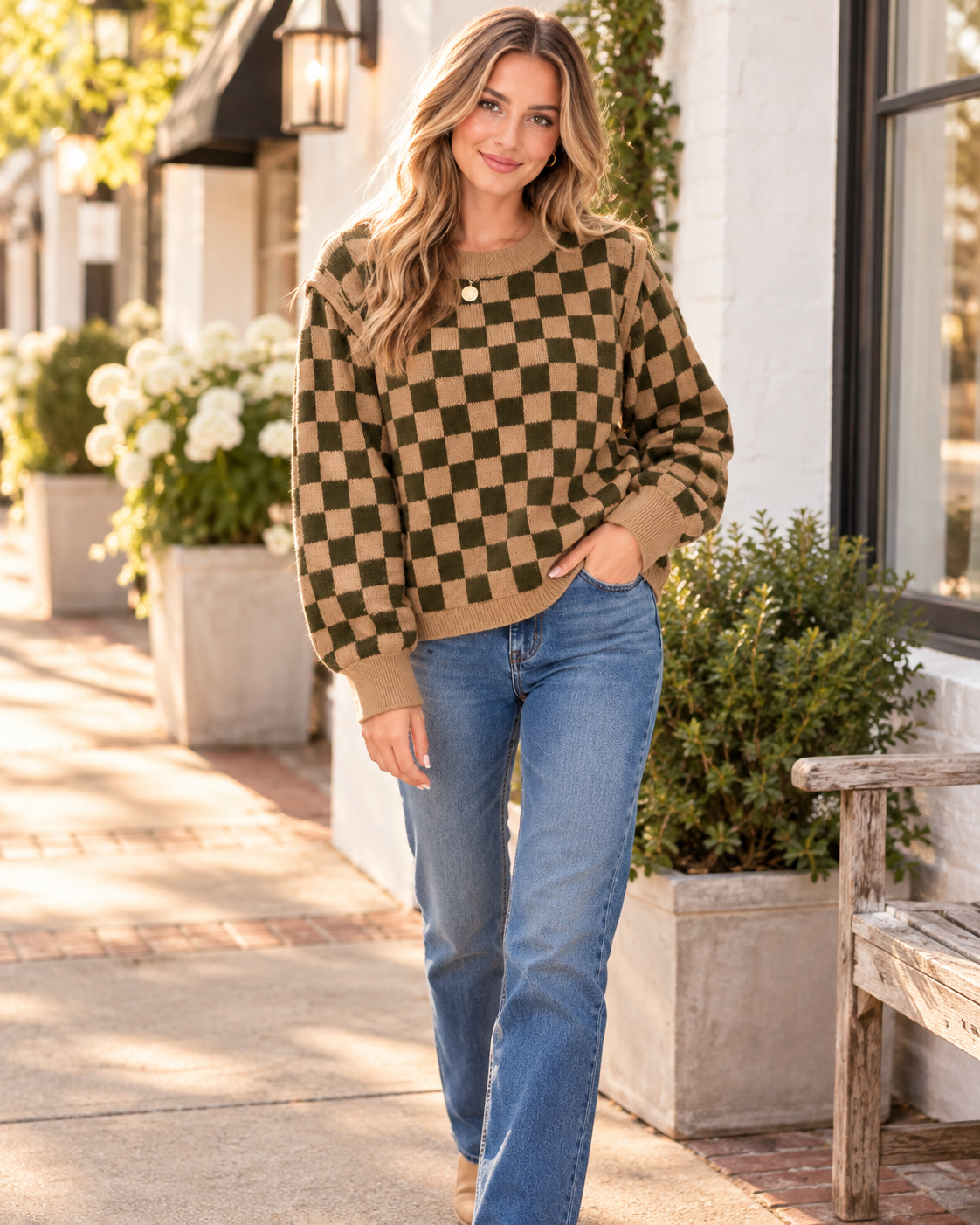 A woman walking outdoors on a sunny sidewalk wearing a brown and tan checkered knit sweater with blue jeans.