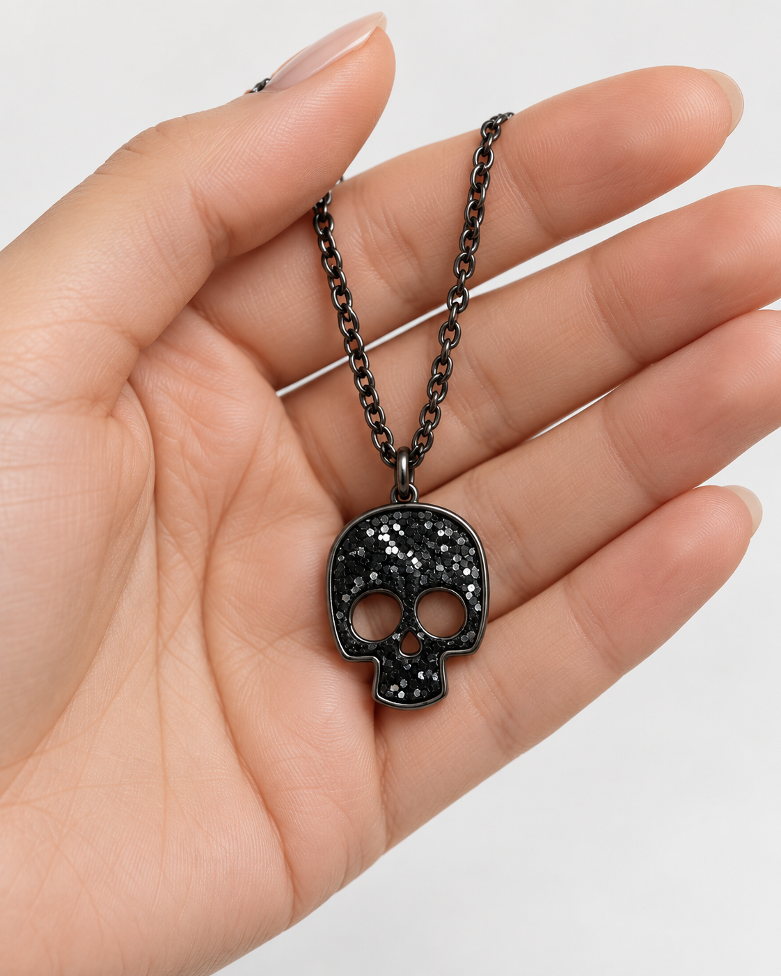 A black skull pendant necklace with a textured, glittery finish, held in a person's hand against a clean white background.