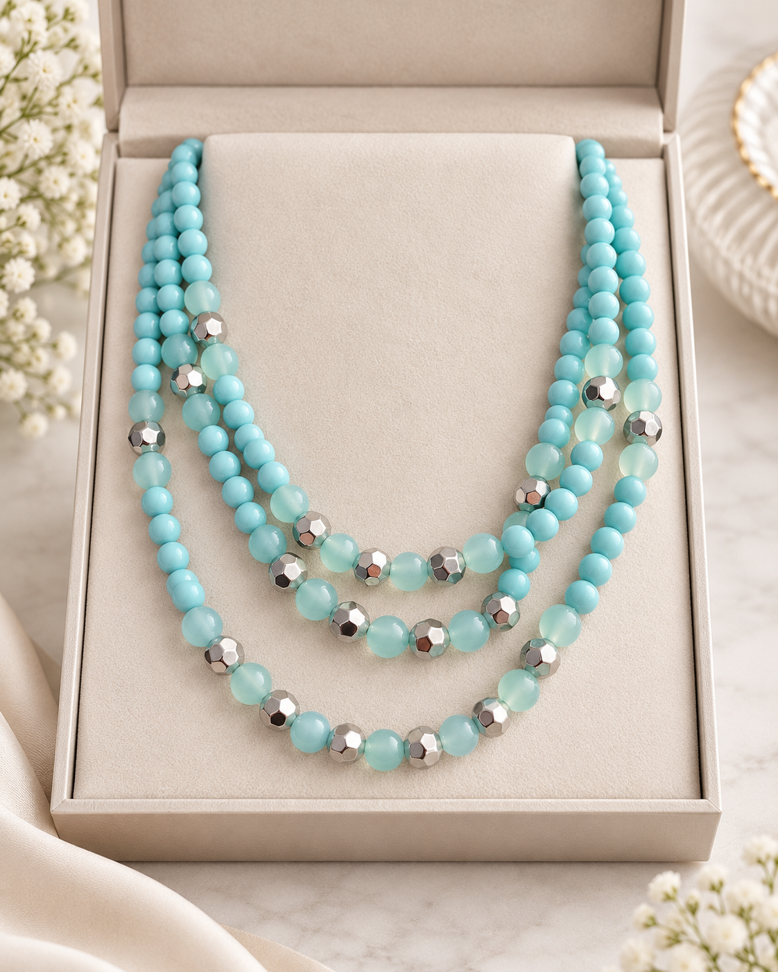 A three-strand beaded necklace in light aqua blue and silver, displayed inside a beige jewelry box on a marble surface with soft fabric and flowers.