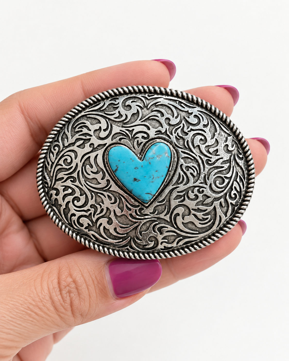 A hand with magenta nail polish holds an oval silver-toned belt buckle featuring intricate scrollwork and a central turquoise heart inlay, set against a plain white background.